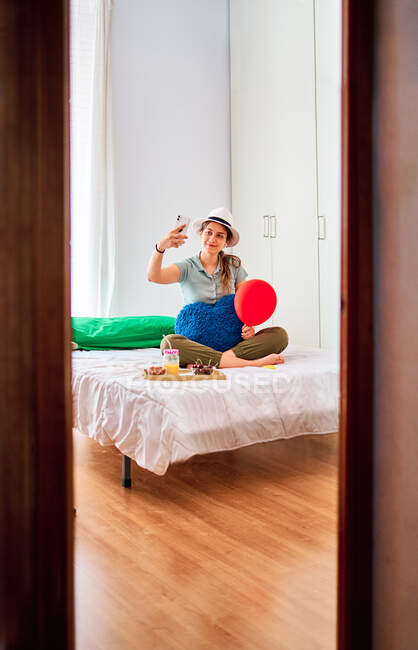 Full length of young female in casual clothes and hat holding red balloon and heart shaped cushion and taking selfie on smartphone while celebrating birthday alone on bed with food and juice — Stock Photo