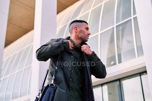 Dal basso dipendente ispanico maschio con valigetta sorridente e guardando la fotocamera mentre camminava sul marciapiede vicino a un edificio moderno in città — Foto stock