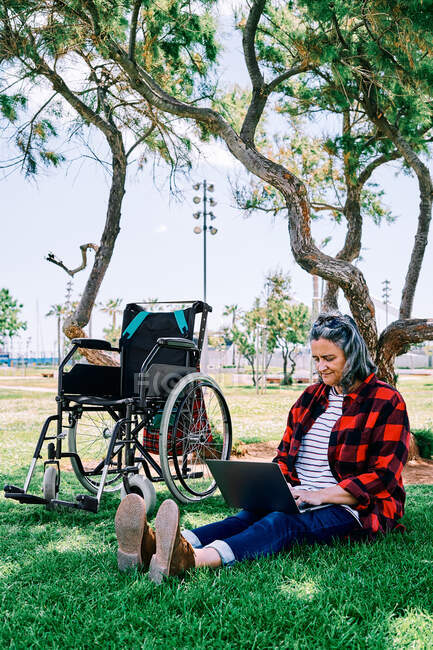Corps complet de femme aux cheveux gris positif avec ordinateur portable sur les genoux assis sur l'herbe verte près de fauteuil roulant dans le parc — Photo de stock