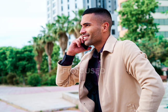 Positive well dressed young Hispanic businessman talking on smartphone and discussing news on urban street with contemporary buildings in background — Stock Photo