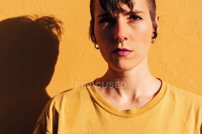 Современная женщина со стильной стрижкой и пирсингом, стоя возле желтой стены в солнечный день на улице — стоковое фото
