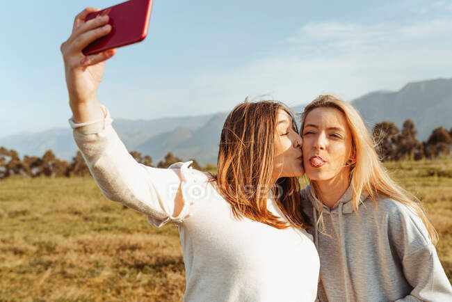 Cheerful young girlfriends taking self photo with smartphone and kissing in countryside of mountains — Stock Photo