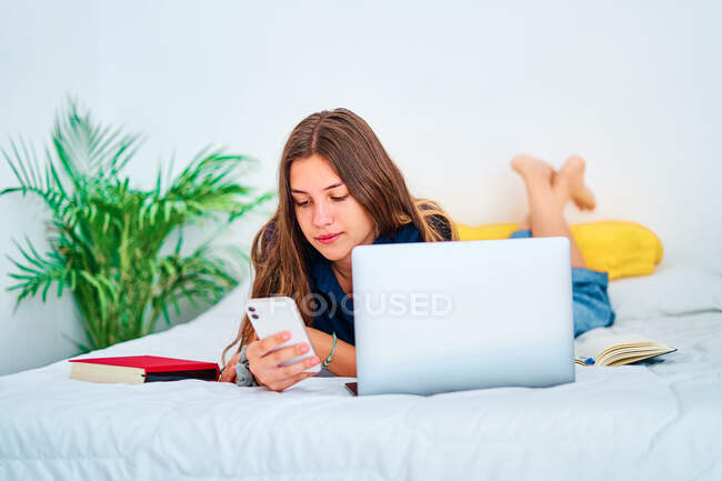 Молода студентка лежить на ліжку з ноутбуком і підручниками та повідомленнями на смартфоні під час дистанційних онлайн-навчань вдома — стокове фото