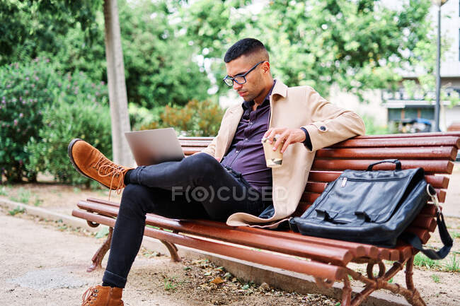 Орієнтований молодий бізнесмен в стильному вбранні і окулярах з чашкою кави, що працює на ноутбуці, сидячи на лавці в міському парку — стокове фото