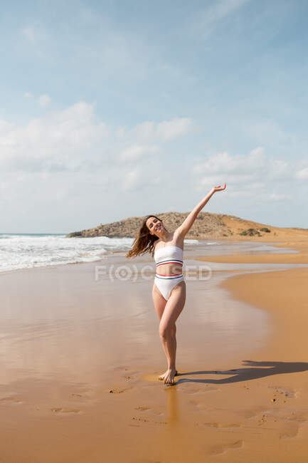 Jeune femme souriante en maillot de bain debout sur une plage de sable regardant la caméra près de l'océan mousseux sous le ciel bleu en plein jour — Photo de stock