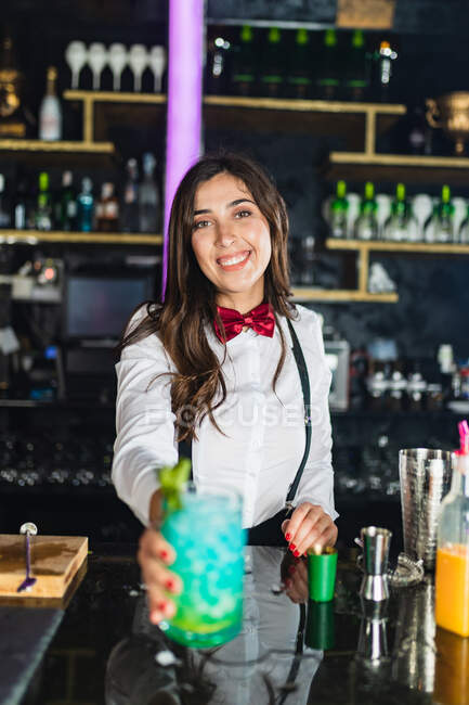 Glückliche Barkeeperin in stylischem Outfit serviert blauen Cocktail mit Minze, während sie an der Theke in einer modernen Bar steht — Stockfoto