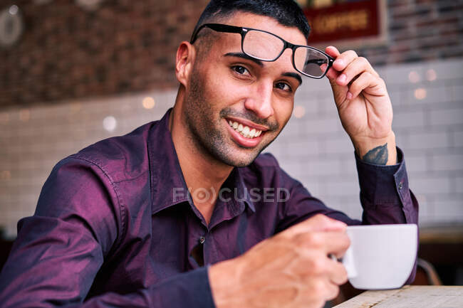 Felice manager maschio ispanico con tazza di bevanda calda togliersi gli occhiali e guardare la fotocamera con sorriso durante la pausa caffè nel caffè — Foto stock