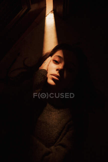 De cima da jovem fêmea silenciosa deitada no chão no quarto escuro com luz brilhando da porta aberta olhando para a câmera — Fotografia de Stock