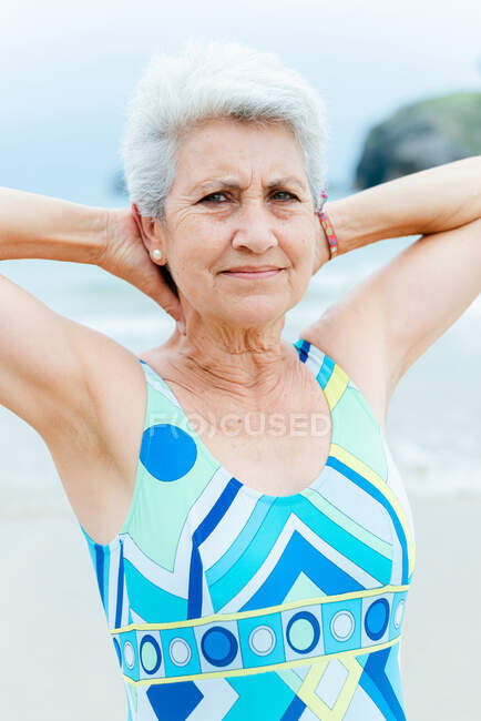 Femme aux cheveux gris vieilli en maillot de bain élégant et lumineux gardant les mains derrière la tête tout en faisant des exercices sur la plage près de la mer — Photo de stock