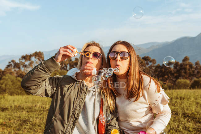 Близкие подруги в солнцезащитных очках дуют мыльные пузыри вместе стоя на лугу в горах — стоковое фото
