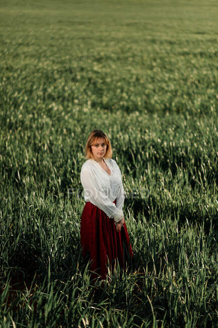 Спокойная молодая женщина в старомодной блузке и юбке, стоящая одна среди высокой зеленой травы в облачный летний день в сельской местности — стоковое фото