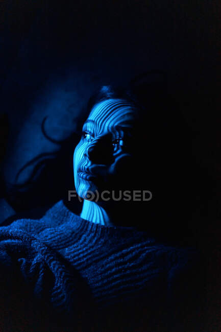 Dreamy young female with light and striped shadow on face looking away pensively while lying in darkness — Stock Photo