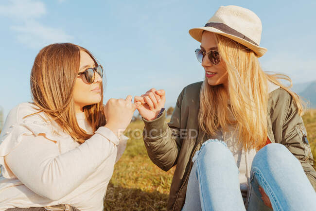 Близкие подружки в шляпе и солнцезащитных очках сидят на лужайке в горах и стучат пальцами при солнечном свете — стоковое фото