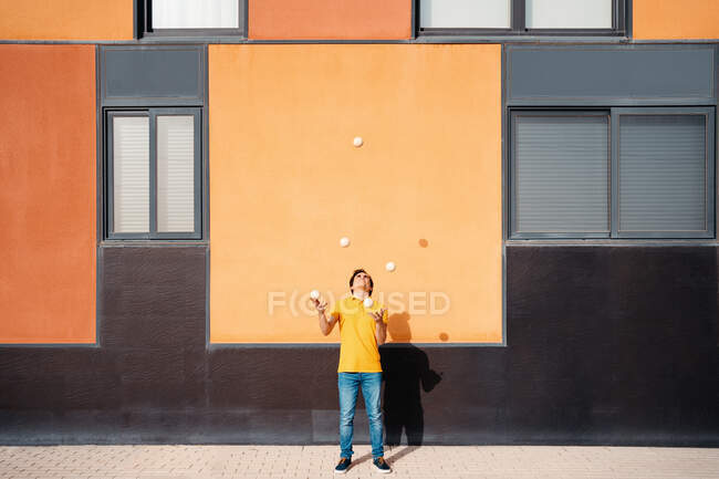Full body of young male performing trick with juggling balls while standing on pavement near bright orange wall — Stock Photo
