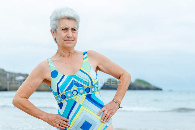 Positive senior female in stylish swimsuit doing side bend exercise while practicing healthy lifestyle and working out on beach against waving ocean — Stock Photo