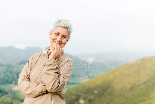 Позитивна літня жінка мандрівник торкається обличчя і дивиться на камеру з посмішкою на розмитому тлі природи — стокове фото