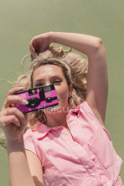 De cima jovem feliz fêmea em vestido rosa tirar fotos na câmera de fotos instantâneas enquanto deitado no chão no dia ensolarado — Fotografia de Stock
