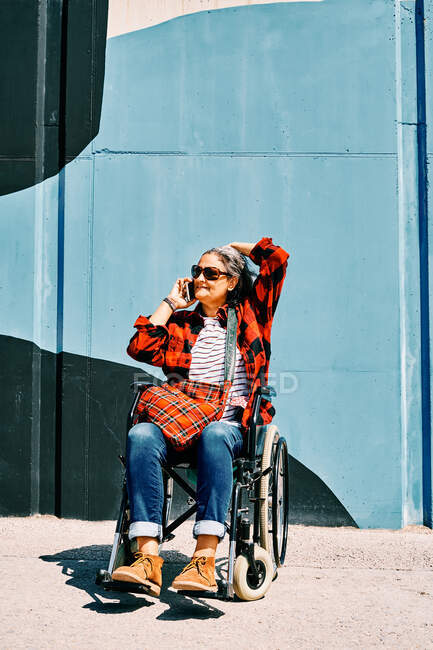 Femme adulte handicapée indépendante en fauteuil roulant parlant sur un téléphone portable près d'un mur peint dans la rue — Photo de stock