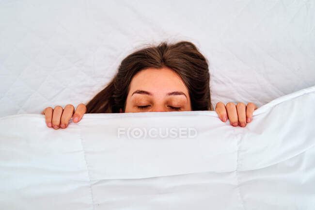 Зверху ледачої молодої жінки, що покриває половину обличчя м'якою білою ковдрою під час завантаження в затишному ліжку вранці — стокове фото