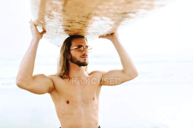 Giovane surfista uomo con i capelli lunghi vestito con muta e occhiali da sole alla moda in piedi portando la tavola da surf sulla testa guardando lontano sulla spiaggia — Foto stock