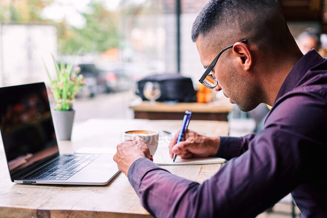 Вид сбоку сфокусированного латиноамериканца с чашкой свежего кофе, сидящего за столом с ноутбуком и делающего заметки в планировщике во время работы в кафе — стоковое фото