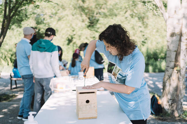 Young female volunteer assembling wooden nesting box for birds during collaboration with activists in green summer park — Stock Photo