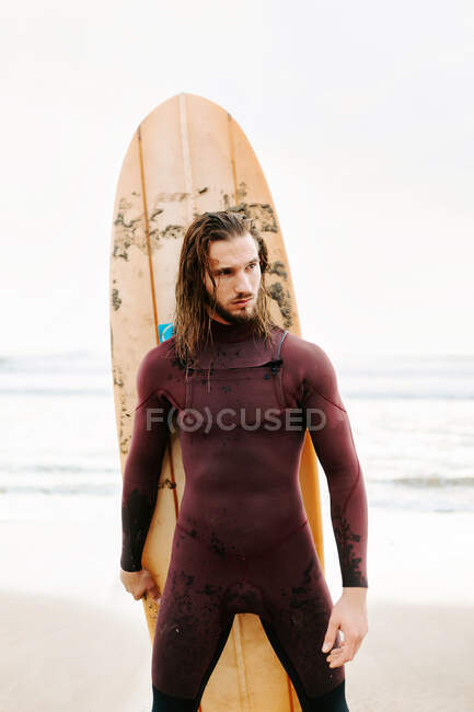 Homem surfista jovem com cabelo comprido vestido de fato de mergulho de pé olhando para longe com prancha de surf em direção à água para pegar uma onda na praia durante o nascer do sol — Fotografia de Stock