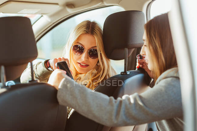 Mulher alegre em óculos de sol voltando para a namorada no banco de trás e compartilhar o telefone enquanto monta carro — Fotografia de Stock
