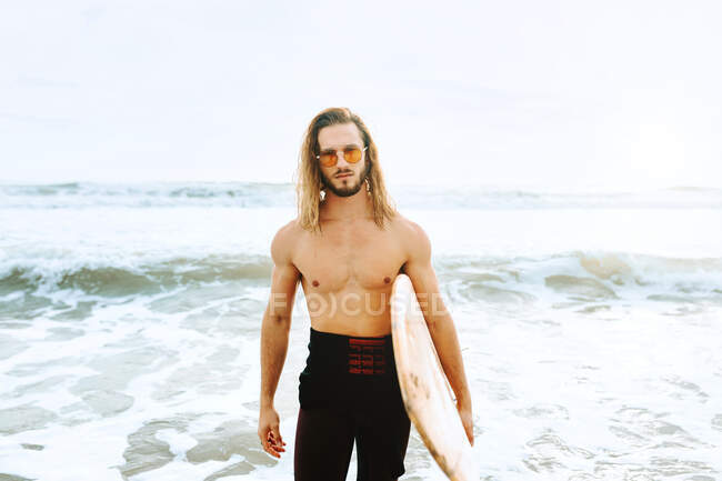 Giovane surfista con i capelli lunghi vestito con muta e occhiali da sole alla moda in piedi guardando la fotocamera con tavola da surf verso l'acqua per catturare un'onda sulla spiaggia — Foto stock
