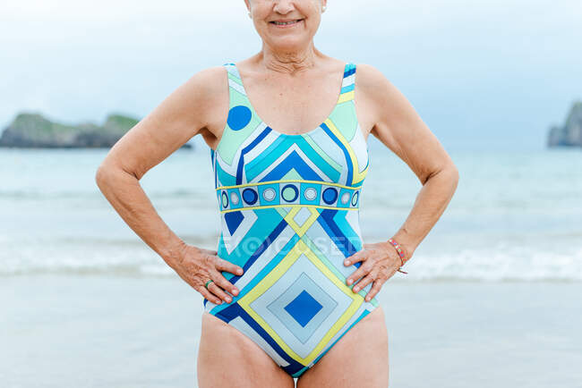 Crop aged female with fit body wearing stylish colorful swimsuit with geometric print standing with hands on waist against sea in summer day — Stock Photo