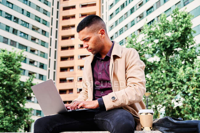 Niedriger Winkel eines fokussierten jungen hispanischen Mannes in stylischer Freizeitkleidung, der ferngesteuert am Laptop an einem freiberuflichen Projekt arbeitet, während er mit einer Tasse Kaffee in der Nähe moderner städtischer Gebäude sitzt — Stockfoto