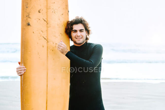 Young surfer man dressed in wetsuit standing looking at camera with surfboard towards the water to catch a wave on the beach during sunrise — Stock Photo