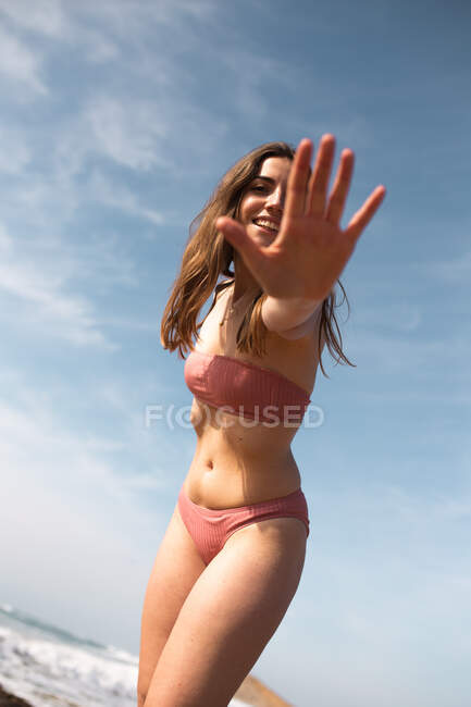 Smiling young female in swimsuit standing on beach while covering face with hand looking at camera on shoreline of ocean — Stock Photo