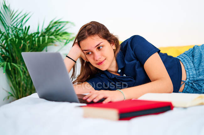 Позитивная молодая женщина-фрилансер в повседневной одежде лежит на кровати и печатает на ноутбуке, работая над удаленным проектом дома — стоковое фото