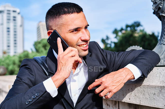 Jeune homme confiant en élégant costume formel parlant sur téléphone portable et souriant tout en se tenant près de la sculpture sur la place urbaine — Photo de stock