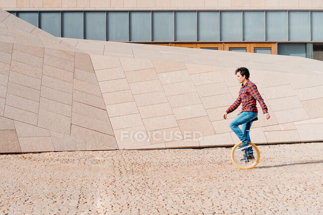 Vista lateral del monociclo masculino contra la construcción contemporánea de piedra de forma geométrica inusual - foto de stock