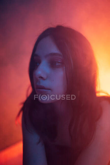 Спокойная молодая модель в платье, сидящая на полу и отводящая взгляд в темную студию с цветными огнями — стоковое фото