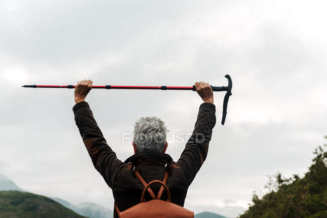 Back view of anonymous aged woman with backpack holding walking stick in raised arms against cloudy gray sky while exploring nature — Stock Photo