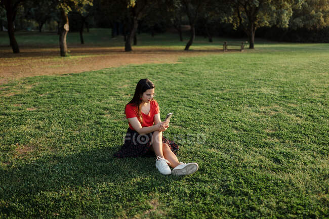Женщина в летней одежде, сидящая на зеленом лугу в парке и серфинг Интернет на мобильном телефоне во время развлечений в выходные дни вечером — стоковое фото
