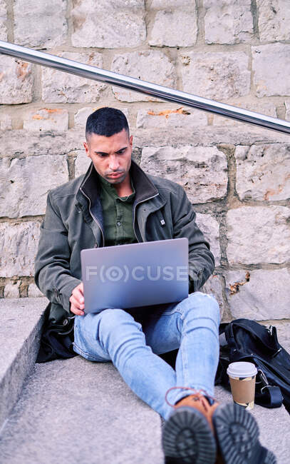 Hispanischer Mann mit Kaffee zum Surfen auf Netbook, während er auf Stufen sitzt und an einem Remote-Projekt in der City Street arbeitet — Stockfoto
