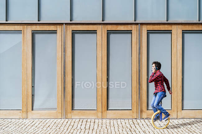 Full body side view of young male in casual outfit answering phone call while riding unicycle on pavement near modern building with geometric facade — Stock Photo