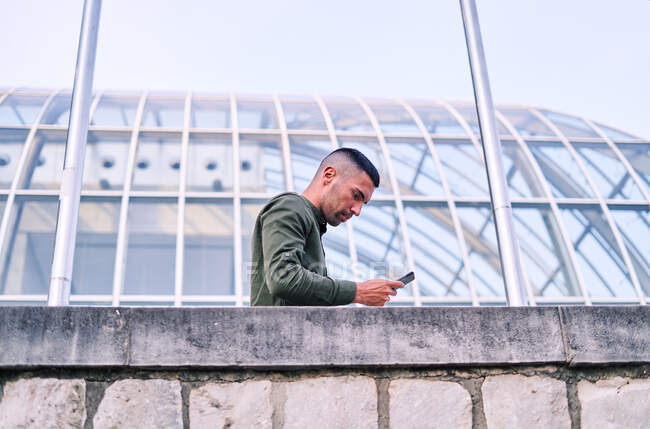 Low angle of adult Hispanic man in shirt leaning on stone wall and browsing cellphone outside contemporary glass building — Stock Photo
