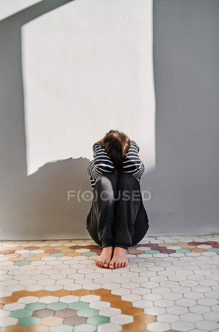 Неузнаваемый несчастный ребенок сидит на полу и закрывает голову руками, страдая от домашнего насилия — стоковое фото