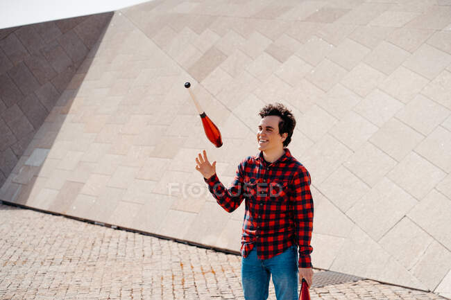 Masculino executando truque com malabarismo clubes enquanto em pé contra contemporânea pedra edifício com incomum geométrica arquitetura — Fotografia de Stock