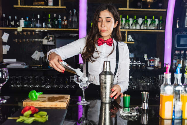 Жінка-бармен в стильному вбранні додає кубики льоду в склянку, готуючи коктейль, що стоїть за стійкою в сучасному барі — стокове фото