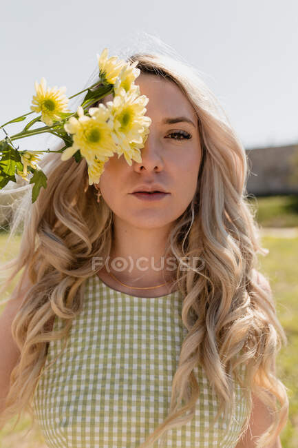 Charming female with long wavy hair hiding face behind blooming flowers while standing in countryside in sunny day — Stock Photo