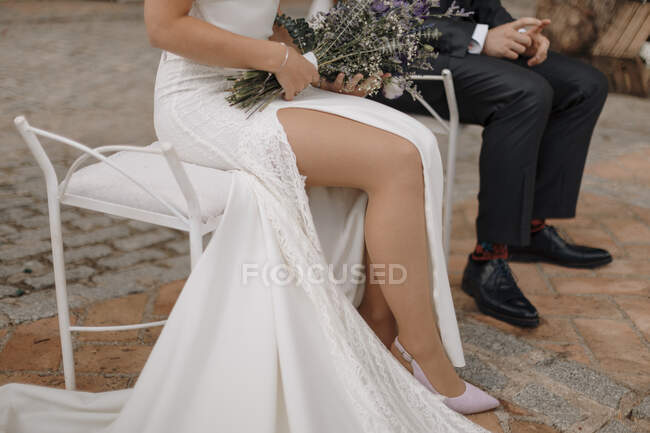 Crop mariée anonyme en robe maxi blanc chic révélant jambe tenant bouquet de fleurs délicates et assis près du marié en costume noir pendant la célébration du mariage — Photo de stock