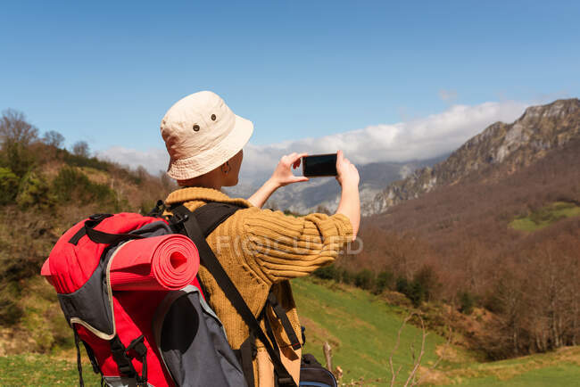 Vista posterior de una mochilera irreconocible tomando fotos de las tierras altas en un teléfono inteligente mientras disfruta de la aventura en verano - foto de stock