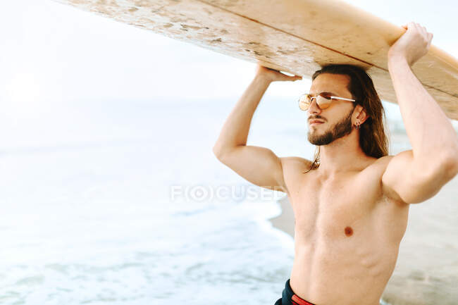 Joven surfista hombre con el pelo largo vestido con traje de neopreno y gafas de sol con estilo de pie llevando la tabla de surf en la cabeza mirando hacia la playa - foto de stock
