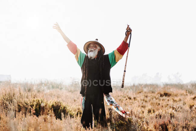Alegre rastafari étnico de edad con rastas mirando hacia otro lado celebrando la victoria mientras está de pie en un prado seco en la naturaleza - foto de stock